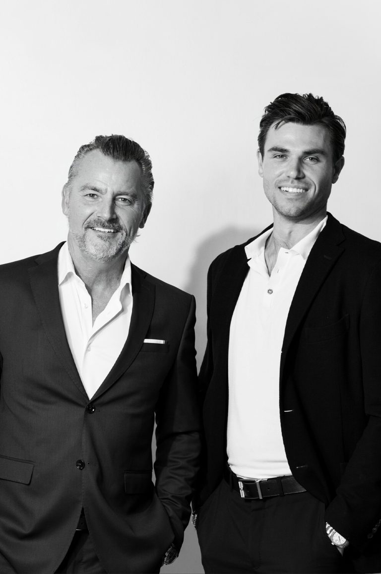 Two men standing next to each other in front of a white wall.