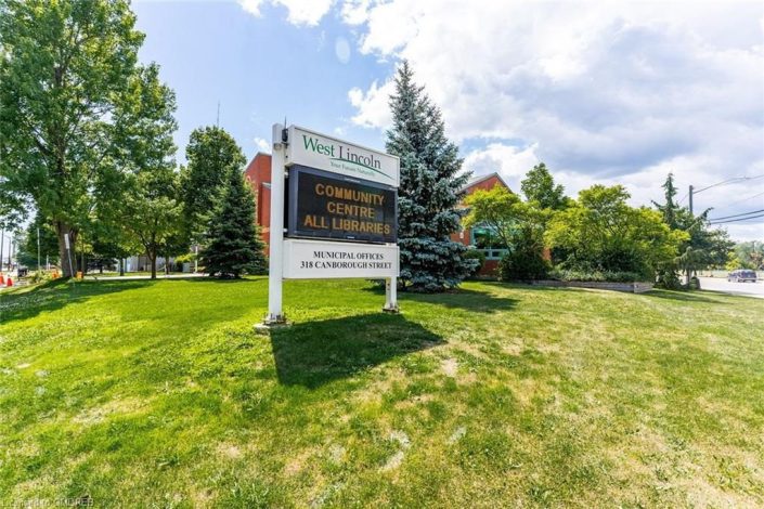 A sign in the grass with trees and buildings behind it.